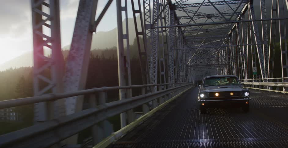 A classic American muscle car drives across a large steel bridge through a scenic mountain landscape during golden hour