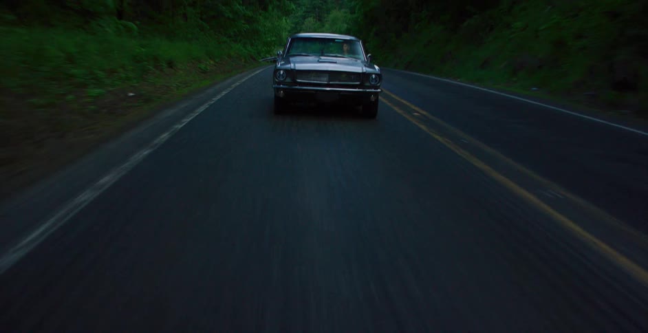 A dynamic shot reveals a classic muscle car driving along a dark, forested two-lane road