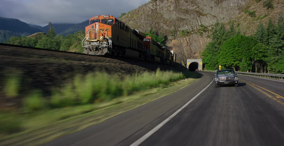 A vintage convertible drives down a scenic highway next to a freight train parked at the base of a rocky mountain