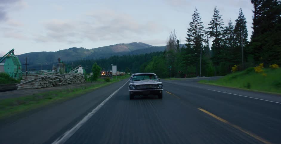 A vintage black muscle car drives down a rural highway past a lumber mill with mountains in the background at dusk