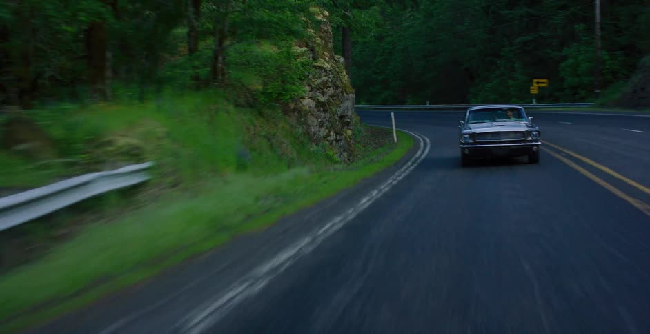 A classic muscle car cruises down a winding two-lane road through a lush green forest in slow motion
