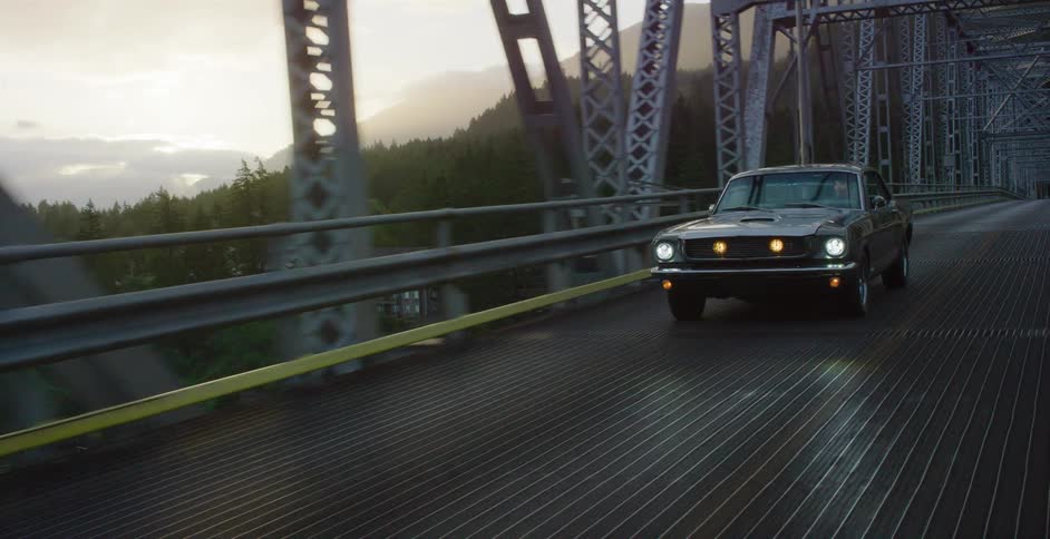 A classic muscle car drives across a scenic metal truss bridge through a mountain forest with its headlights on at dusk