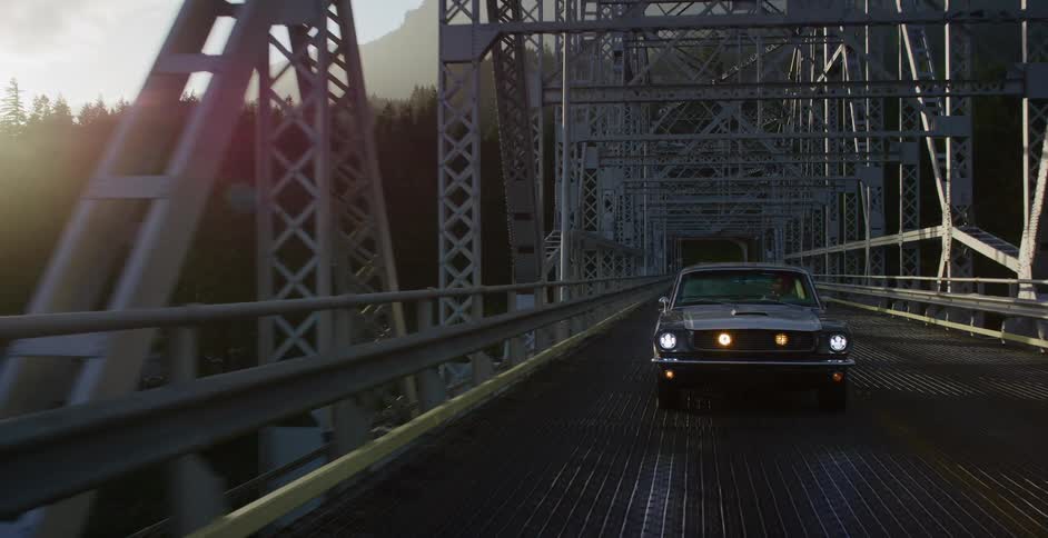 A classic black Ford Mustang with its headlights on drives across a metal truss bridge as the sun sets behind a mountain forest