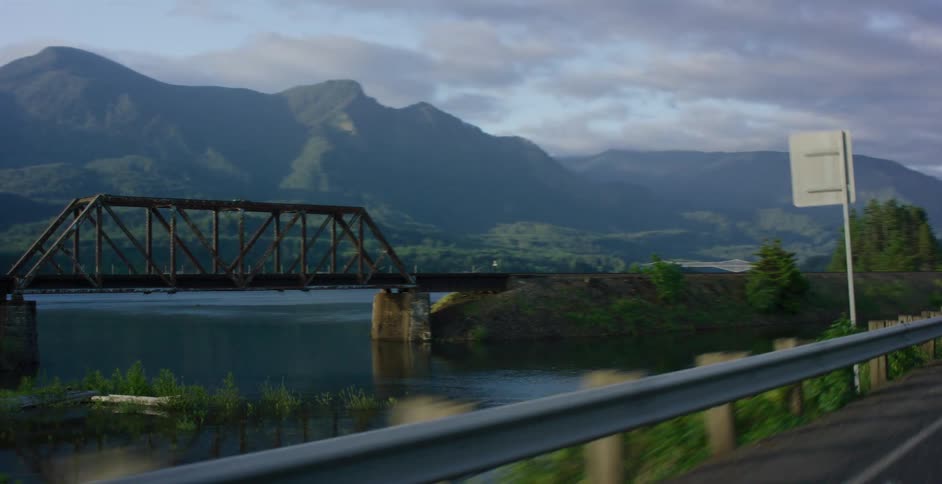 A tracking shot from a car reveals a rustic truss bridge over a river before pulling back to show a classic car driving on a scenic mountain highway