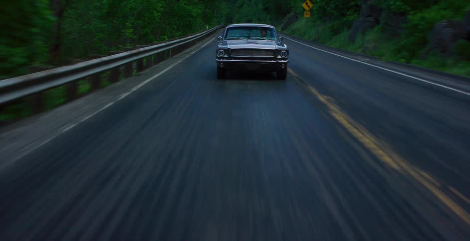A classic dark muscle car speeds down a two-lane road through a dense, moody forest