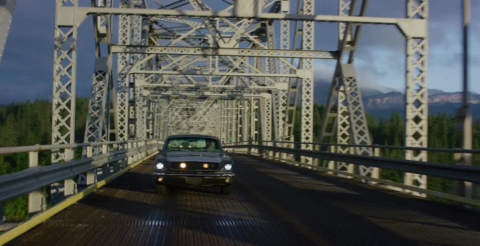 A classic Ford Mustang with headlights on drives across an iconic steel truss bridge surrounded by a scenic forest and mountain landscape