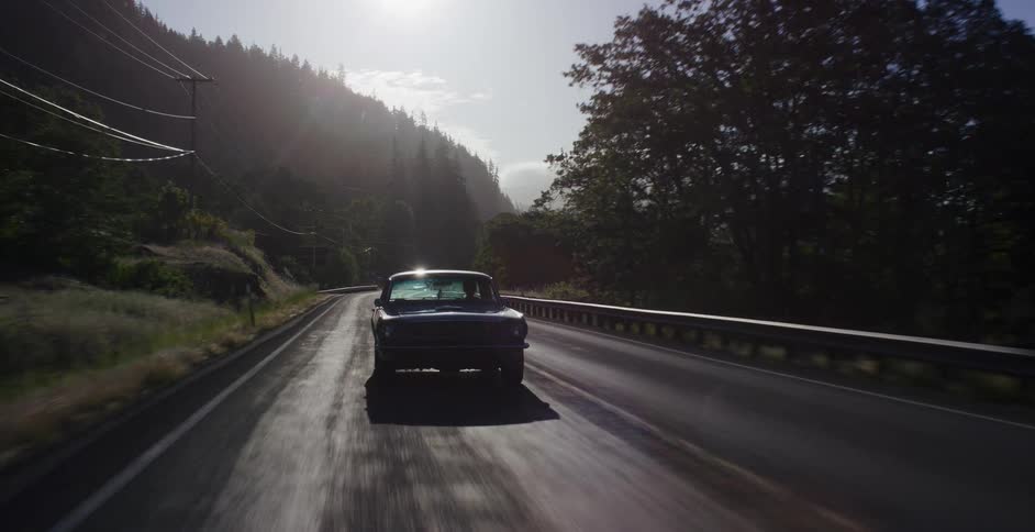 A classic black car drives down a scenic two-lane highway with a bright sun flare in the background