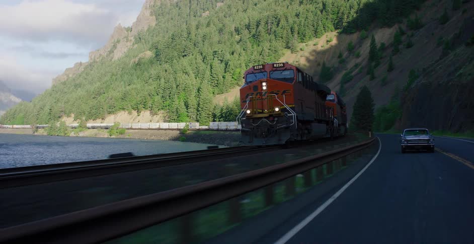 A powerful red freight train races a classic black car along a scenic mountain highway next to a river