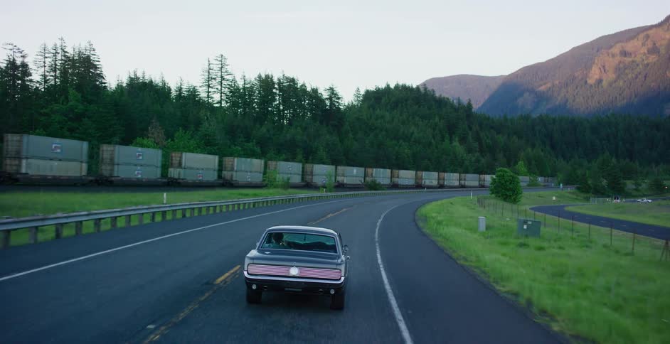 A classic black muscle car cruises down a winding country road at dusk, driving alongside a long freight train with forested mountains in the background