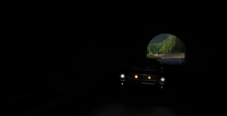 A classic silver muscle car drives along a picturesque highway next to a river and mountains, passing through a tunnel on a beautiful day