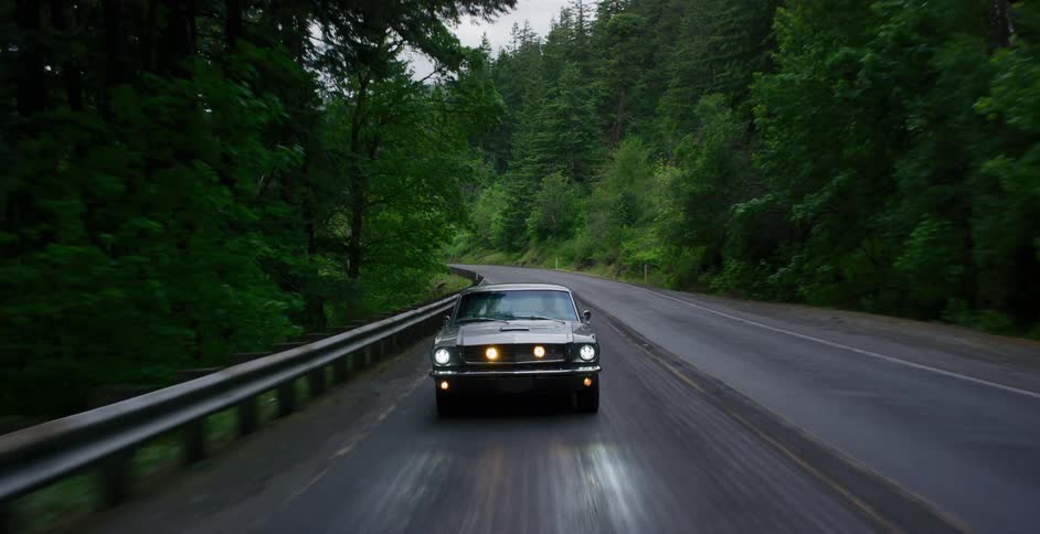 A cinematic front-facing shot follows a classic silver muscle car as it drives down a wet, winding road through a lush, dark green forest