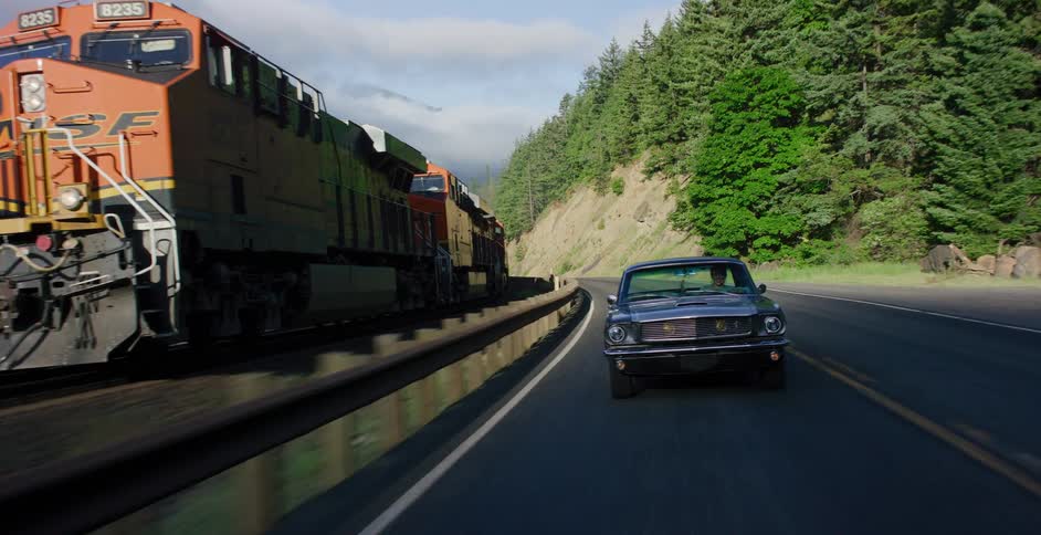 A classic black Mustang drives down a scenic highway alongside a fast-moving freight train in the mountains
