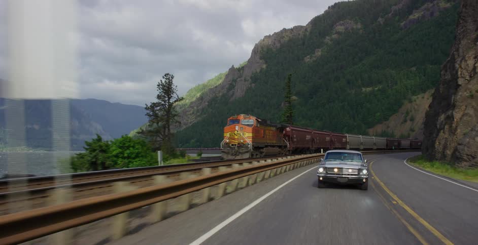 A classic muscle car drives down a winding highway next to a long freight train traveling through a scenic mountain gorge