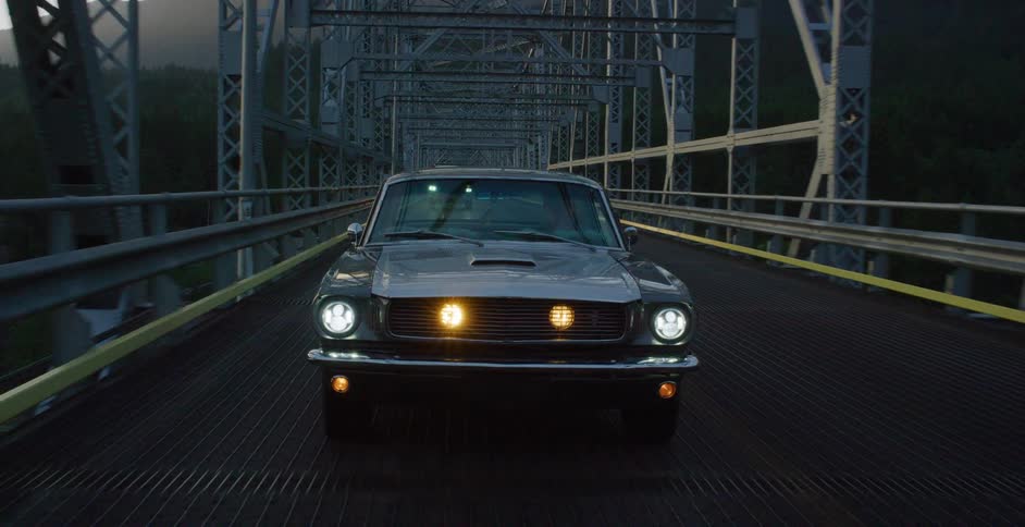 A classic black Ford Mustang drives across a scenic steel truss bridge over a river in the mountains at dusk