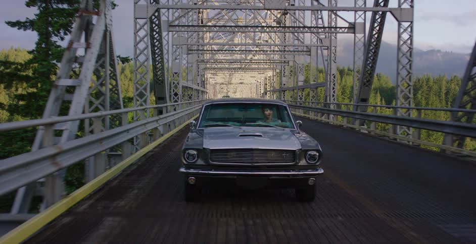 A vintage silver Mustang drives across a long metal truss bridge through a lush green forest