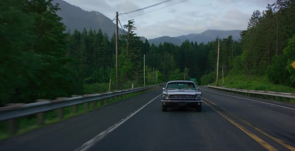 A silver classic car drives steadily toward the camera on a two-lane road winding through a lush, green forest with mountains in the background