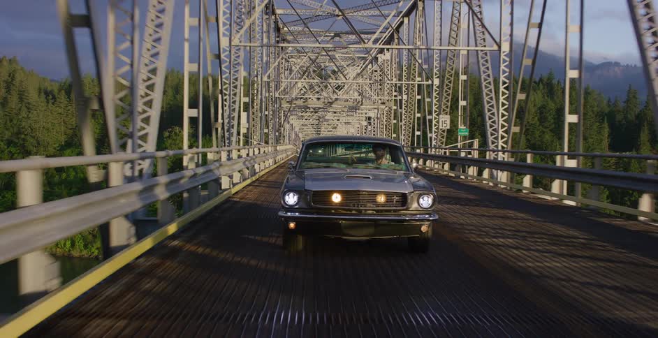 A classic silver Ford Mustang drives across a scenic metal truss bridge over a river surrounded by lush forests and mountains