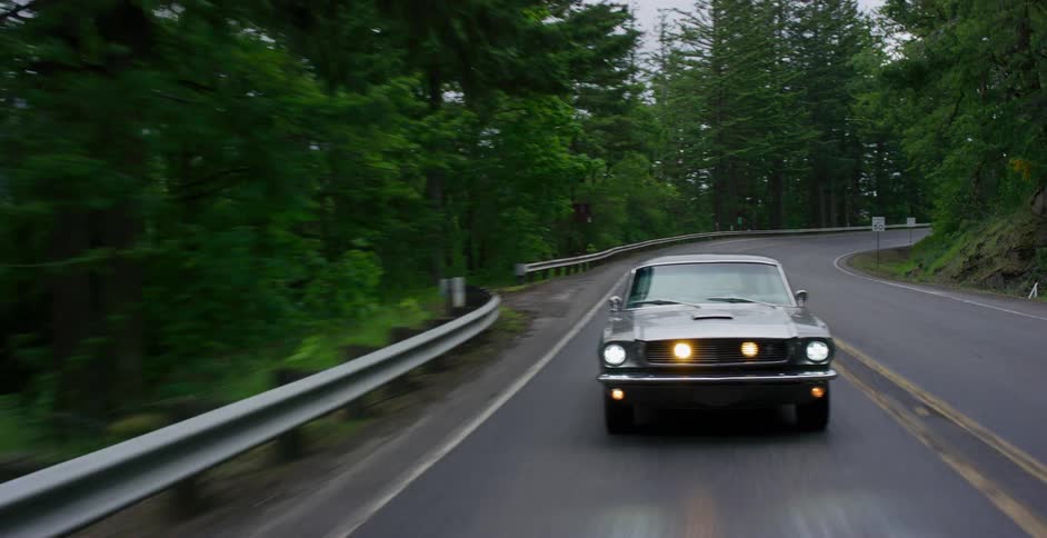 A classic silver Ford Mustang with its headlights on drives along a winding, tree-lined road through a lush forest on an overcast day