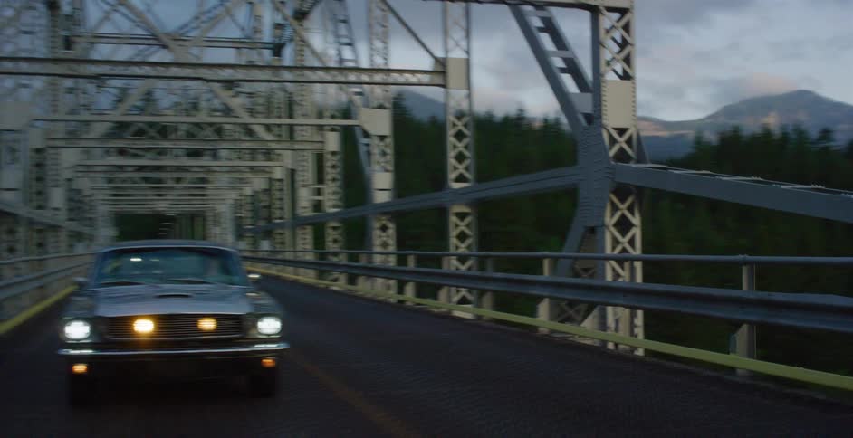 A classic Ford Mustang with its headlights on drives across a long steel truss bridge over a river at dusk