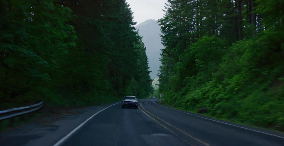 Reverse dolly shot of a classic car driving on a remote road surrounded by a dense forest and misty mountains in the distance
