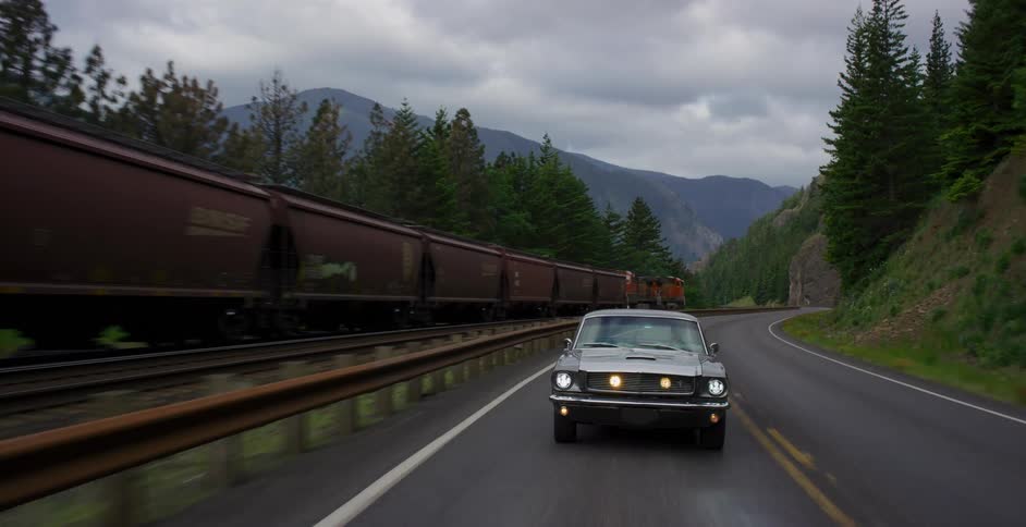 A classic grey Ford Mustang drives on a wet two-lane road through a mountain pass as a long freight train passes on parallel tracks