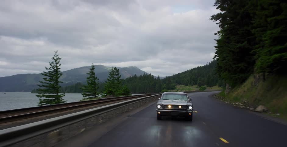 A classic muscle car cruises down a scenic highway alongside a large lake with mountains in the background under an overcast sky