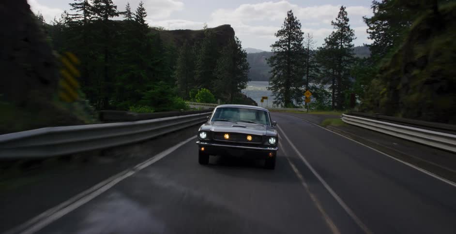 A classic silver muscle car drives along a winding road through a beautiful, forested mountain landscape