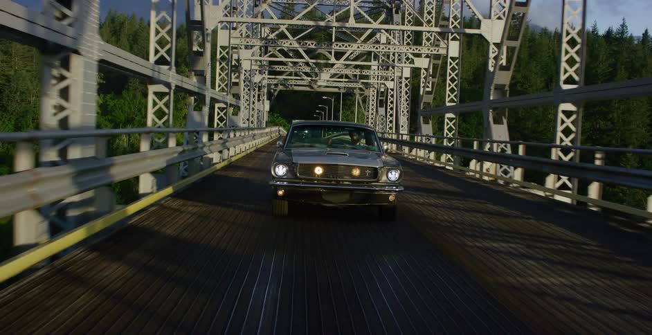 Camera tilts down the steel trusses of a bridge to reveal a classic Ford Mustang driving towards the viewer