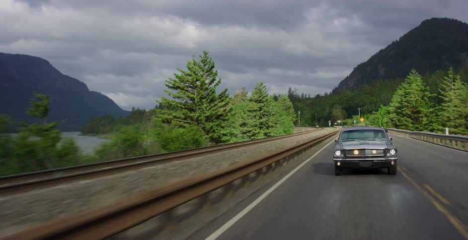 Tracking shot of a classic grey Ford Mustang cruising down a scenic highway next to a river and mountains under a cloudy sky