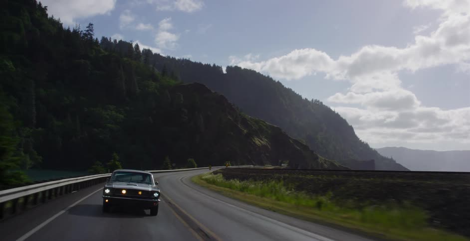 A vintage muscle car drives along a scenic, winding mountain road and into a dark tunnel