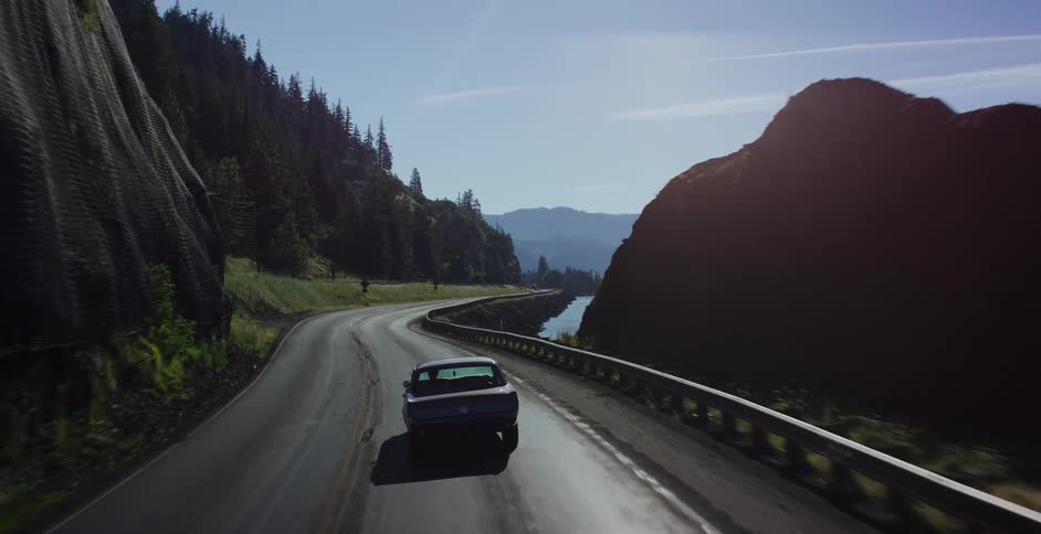 Following a vintage muscle car as it emerges from a dark tunnel onto a sunlit, winding highway along a scenic river