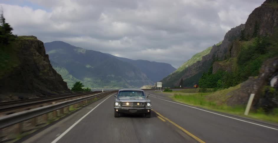 A classic grey Ford Mustang cruises down a scenic highway through a mountainous gorge next to a river on an overcast day