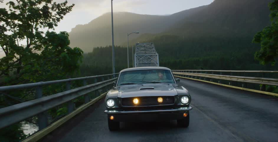 A classic muscle car drives along a scenic bridge surrounded by misty mountains and lush green forests at sunrise