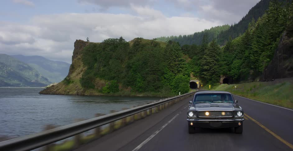 A classic gray muscle car cruises down a scenic highway alongside a vast river, with forested mountains and tunnels in the distance