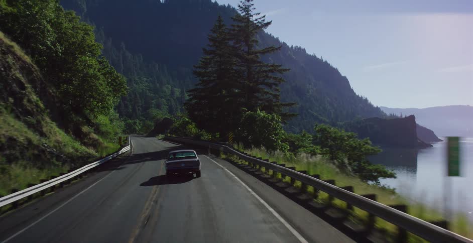 A point-of-view shot follows a vintage car as it exits a dark tunnel and travels along a scenic mountain road next to a river on a sunny day