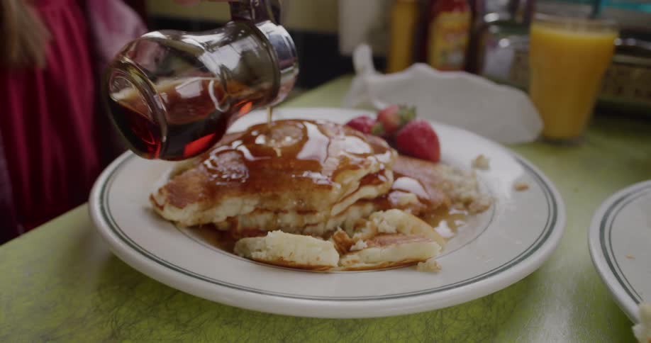 A young girl pours maple syrup over a stack of fluffy pancakes with strawberries at a diner