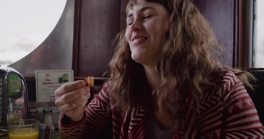 Slow motion close-up of a happy woman with brown hair laughing while eating breakfast in a retro diner booth