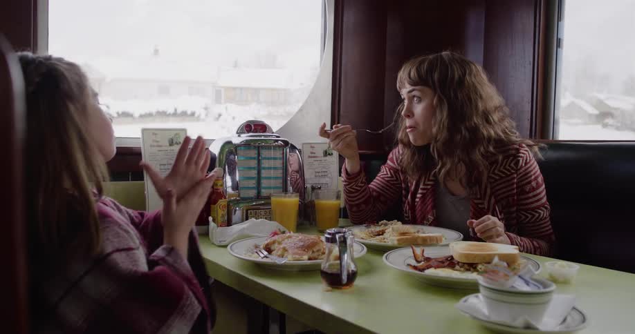 A mother and daughter share a conversation while eating breakfast in a cozy diner booth on a snowy day in slow motion