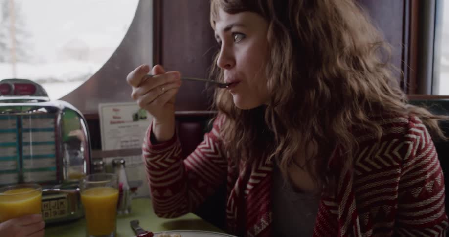 A young woman eats a bite of her breakfast in slow motion while sitting in a classic diner booth on a snowy day