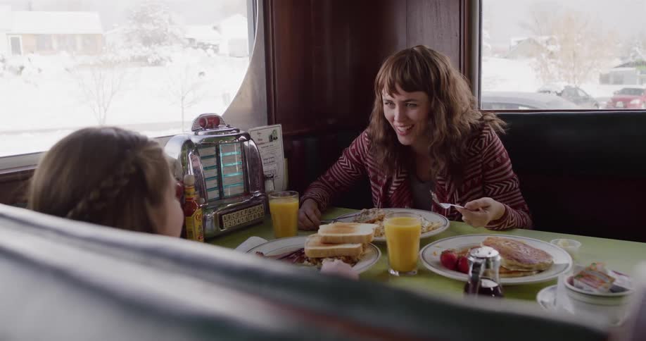 A happy mother and daughter laugh while eating breakfast in a cozy diner on a snowy day