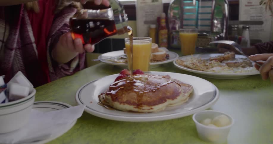 Slow motion tilt down from a smiling girl as she pours syrup over a stack of pancakes at a retro diner with her mom