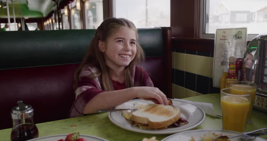 A smiling young girl sits in a retro diner booth, happily anticipating her breakfast on a cold winter morning