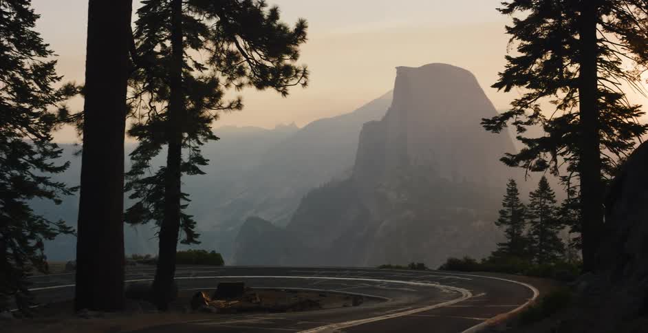 A winding mountain road through a pine forest reveals a majestic view of Half Dome at sunrise in Yosemite National Park