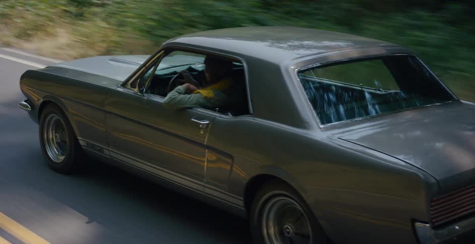 Side tracking shot of a man driving a vintage silver Ford Mustang down a scenic country road