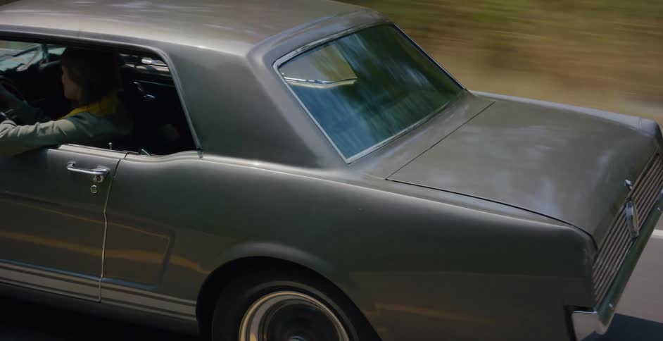 A person cruises in a classic silver Ford Mustang along a scenic, tree-lined highway on a sunny day