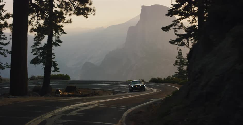 A classic Ford Mustang drives around a scenic mountain curve revealing an epic, hazy sunrise over Yosemite Valley and Half Dome