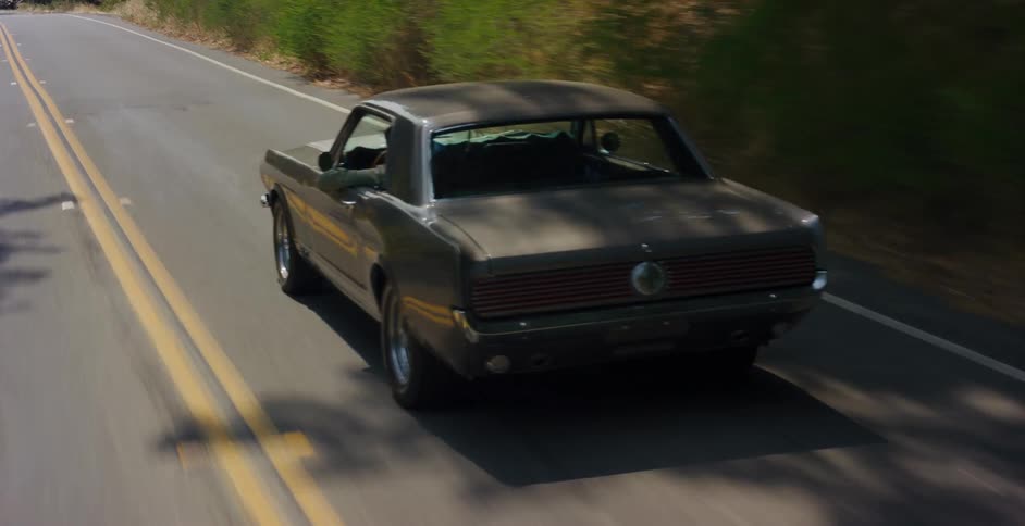 A classic silver Ford Mustang cruises down a sunlit two-lane country road