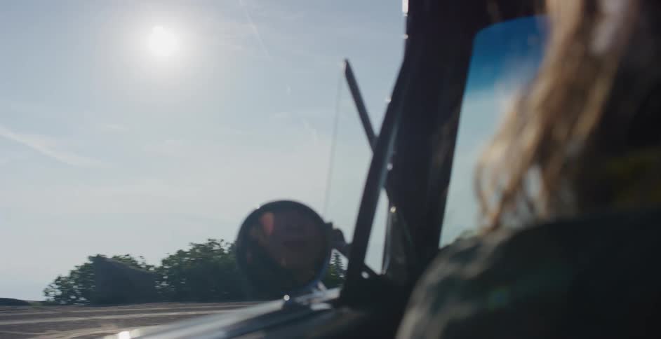 A happy woman's face is reflected in the side-view mirror of a classic car as she drives along a sunny, scenic road