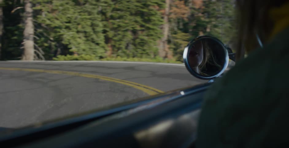 The reflection of a young woman driving is seen in the side mirror of her vintage car on a curvy mountain road