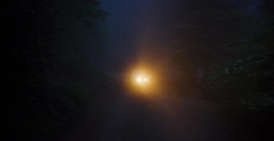 Aerial shot pulls back from a car's glowing headlights on a spooky, foggy forest road at night, tilting up to reveal the towering trees